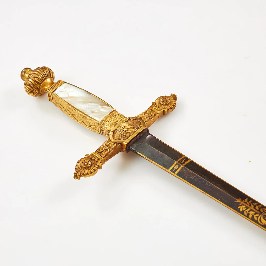 ÄMBETSMANNAVÄRJA. Empire, klinga med blånering och förgylld dekor i form av bla. Karl XIV Johans monogram, korsformigt mässingsfäste med kavelplattor i pärlemor, balja i svart läder med två fögyllda beslag. — Edged weapons — Stockholms Auktionsverk Sickla