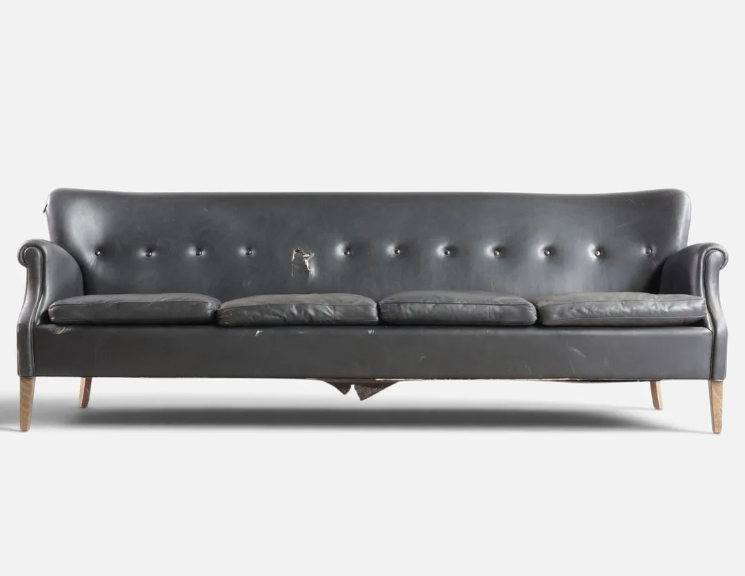 Fritstående fire-pers. sofa med skammel, betrukket med knapsyet sort skind, tilspidsede ben af bøg. Antageligvis udført hos Fritz Hansen. Danmark, 1940-50'erne. — Sofas & Seatings — Woxholt Auktioner